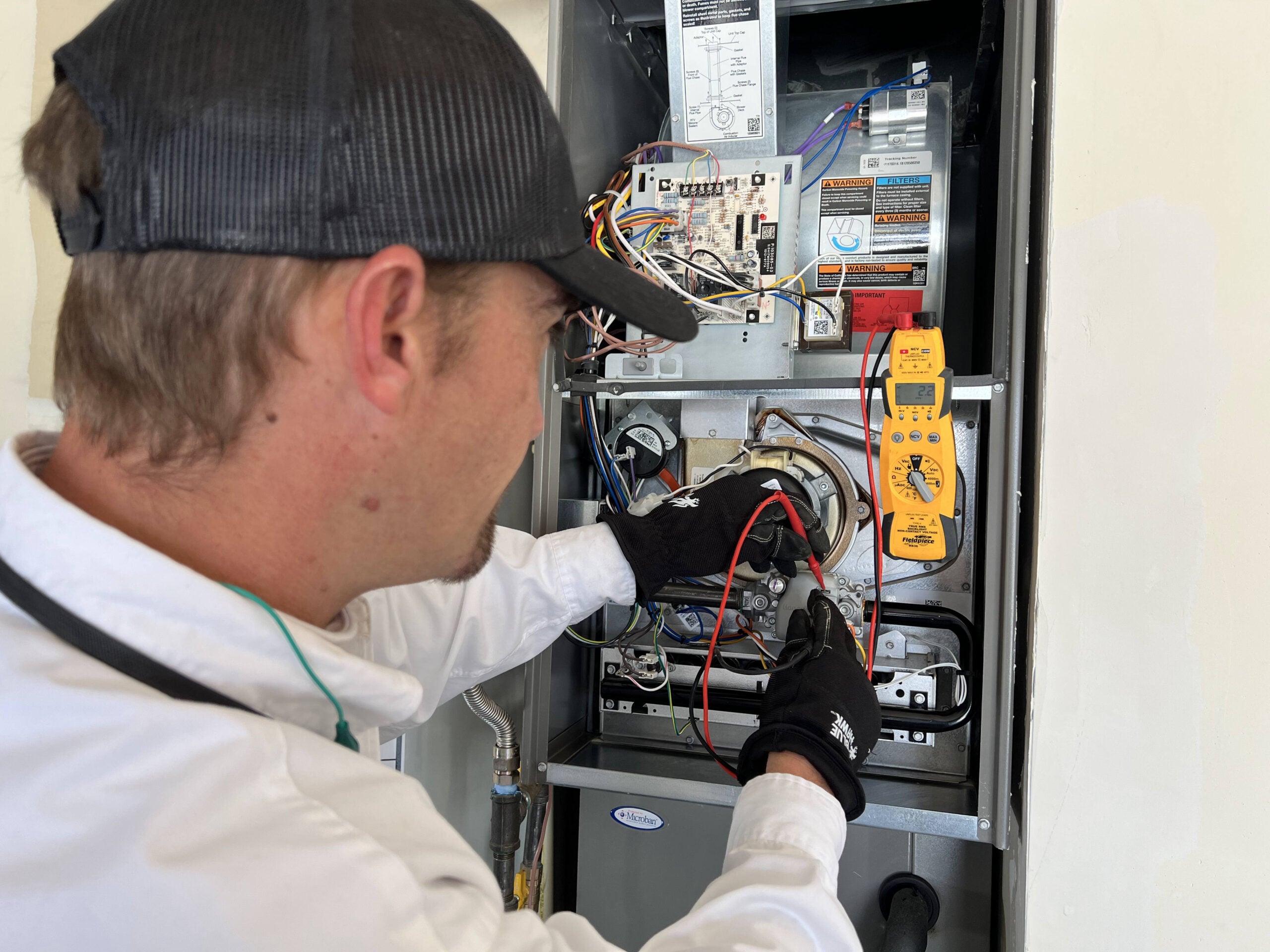 Technician in cap and gloves examines furnace internal components with yellow digital multimeter during maintenance inspection.