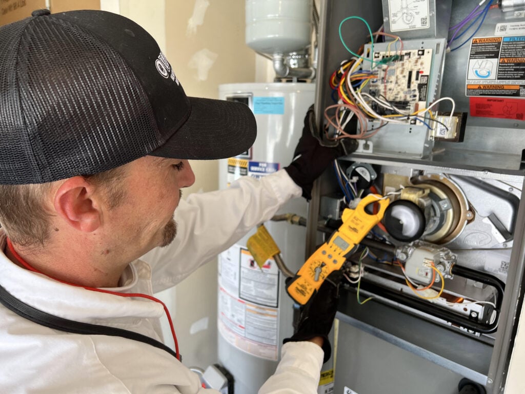 Technician in black cap using yellow clamp meter to test electrical connections inside furnace unit with visible wiring and components.