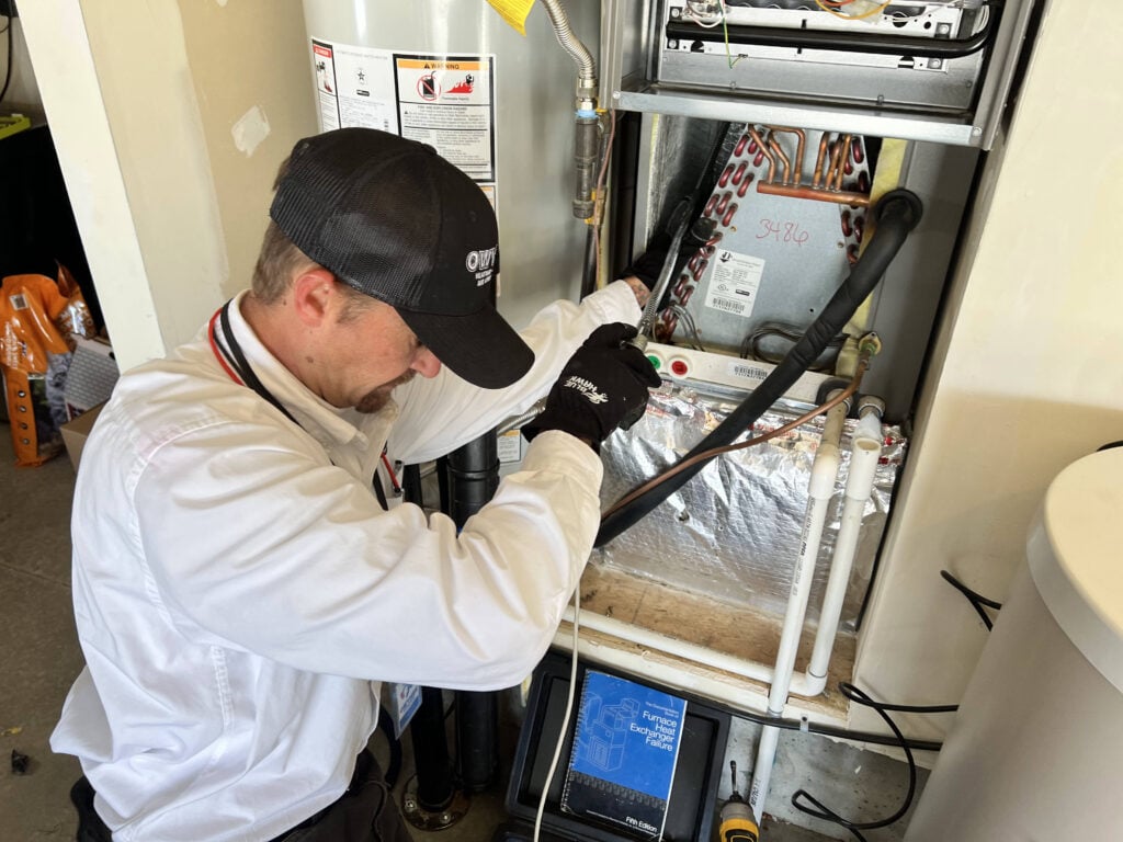 Technician in white shirt and black cap wearing gloves performs maintenance on an indoor furnace unit with visible copper piping and control board.