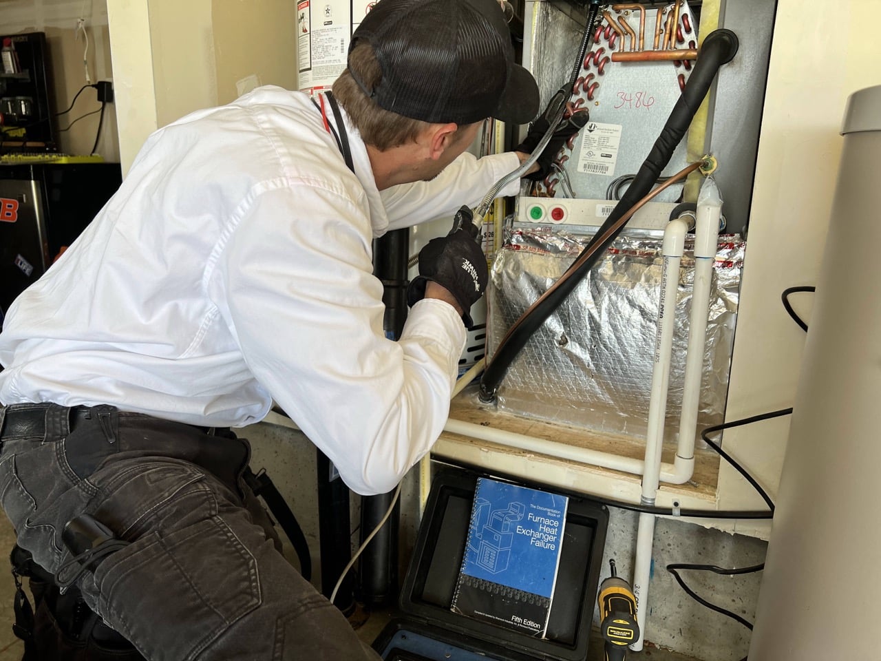 Technician in white jacket examining furnace heat exchanger with diagnostic tools and reference manual visible.