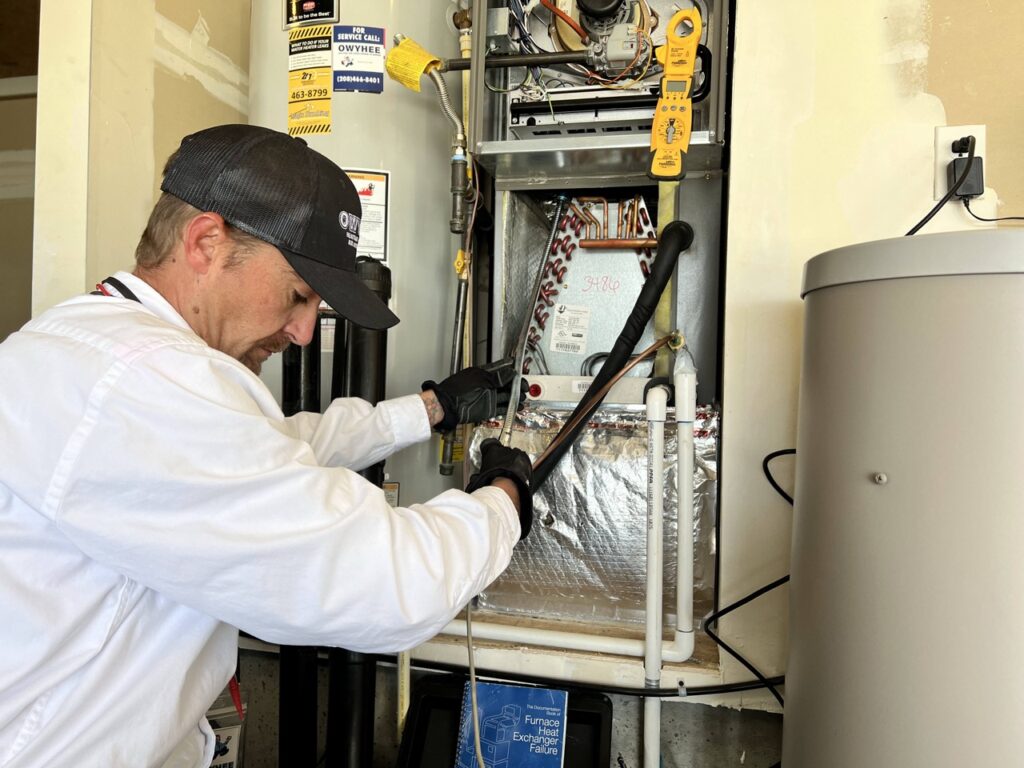 HVAC technician in white jacket inspecting furnace heat exchanger with tools. Service call tag visible on wall.