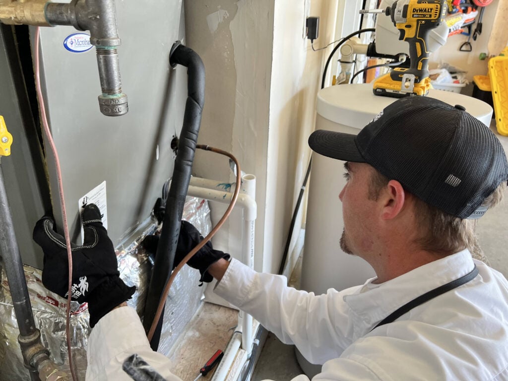 Technician in black cap and white shirt installs copper piping on water heater unit in utility room.