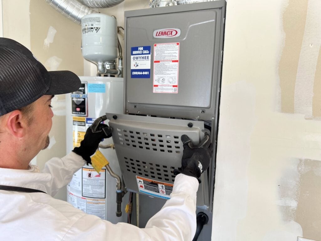 Technician in black gloves and white coveralls installs a gray Lennox furnace unit with service information visible.
