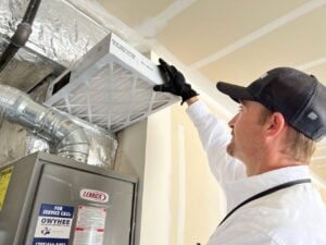 HVAC technician in white shirt and black cap installing a pleated air filter into a Lennox furnace unit mounted on ceiling.