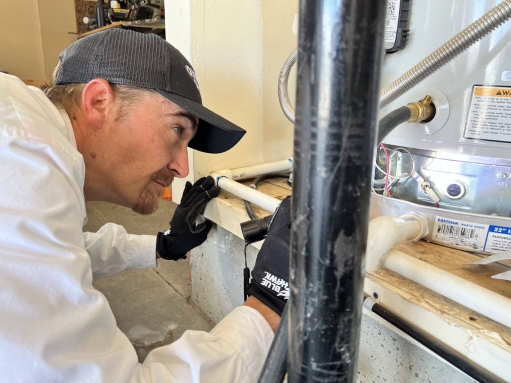 Plumber in black cap and white shirt installing white PVC pipes on a water heater system indoors.