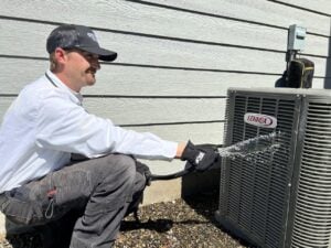 HVAC technician in white shirt and black cap crouches beside a Lennox outdoor air conditioning unit against a gray siding house.