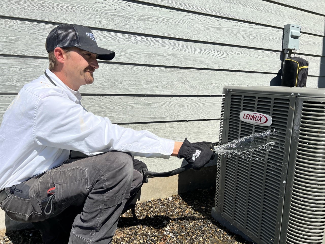 HVAC technician in white shirt and black cap crouches beside a Lennox outdoor air conditioning unit against a gray siding house.