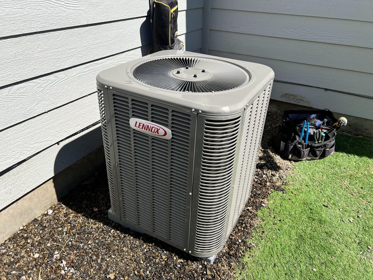 Lennox central air conditioning condenser unit installed against white vinyl siding with service equipment nearby.