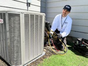 Professional technician in white shirt and black cap performs maintenance on residential air conditioning unit with tools and gauges.