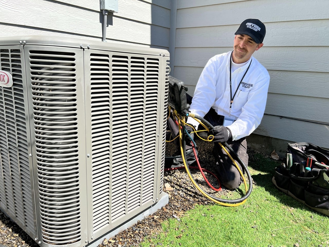 Professional technician in white shirt and black cap performs maintenance on residential air conditioning unit with tools and gauges.