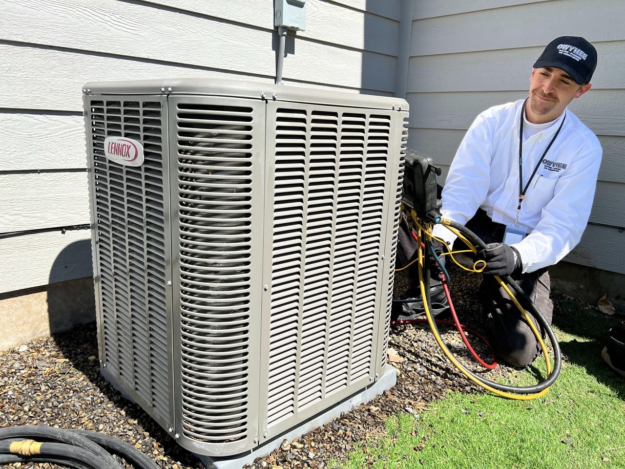 HVAC technician in white shirt and cap performs maintenance on outdoor Lennox air conditioning unit with diagnostic equipment and hoses.