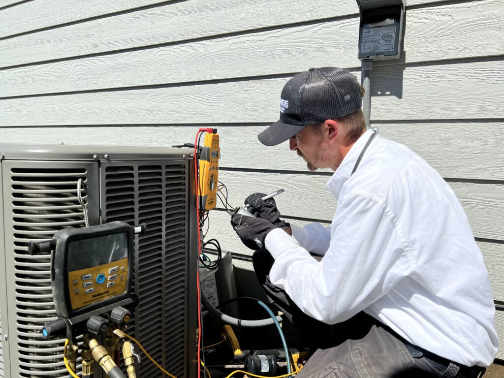 Technician in white jacket and black cap using diagnostic equipment to service an outdoor air conditioning unit mounted on a house.