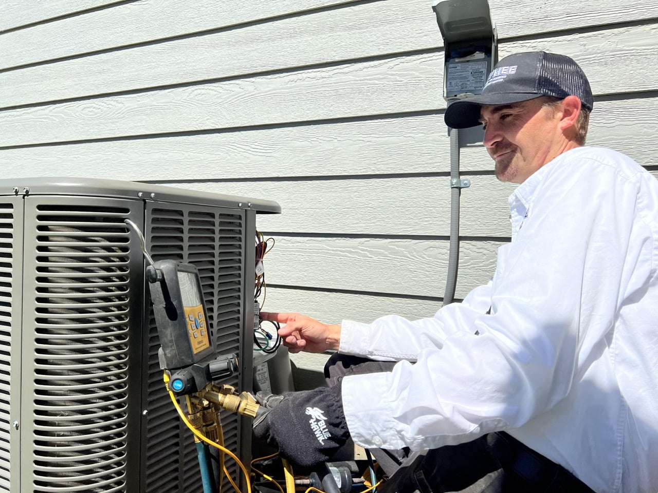 Technician in white shirt and cap using digital gauge meter to service outdoor air conditioning unit against light gray siding.
