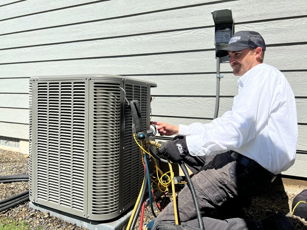 Technician in white shirt and cap servicing an outdoor air conditioning unit with diagnostic tools and colored wires visible.