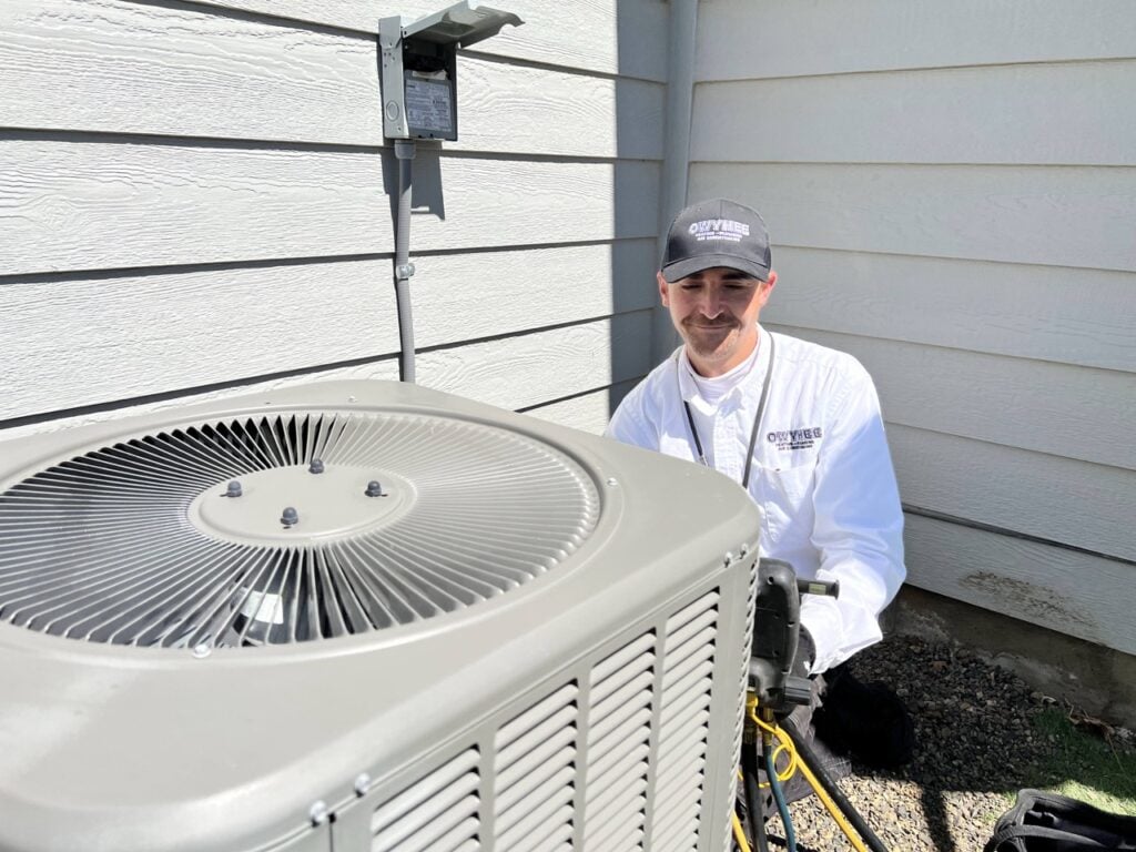 Technician in white shirt and cap kneels beside a large white air conditioning condenser unit against a house with horizontal siding.