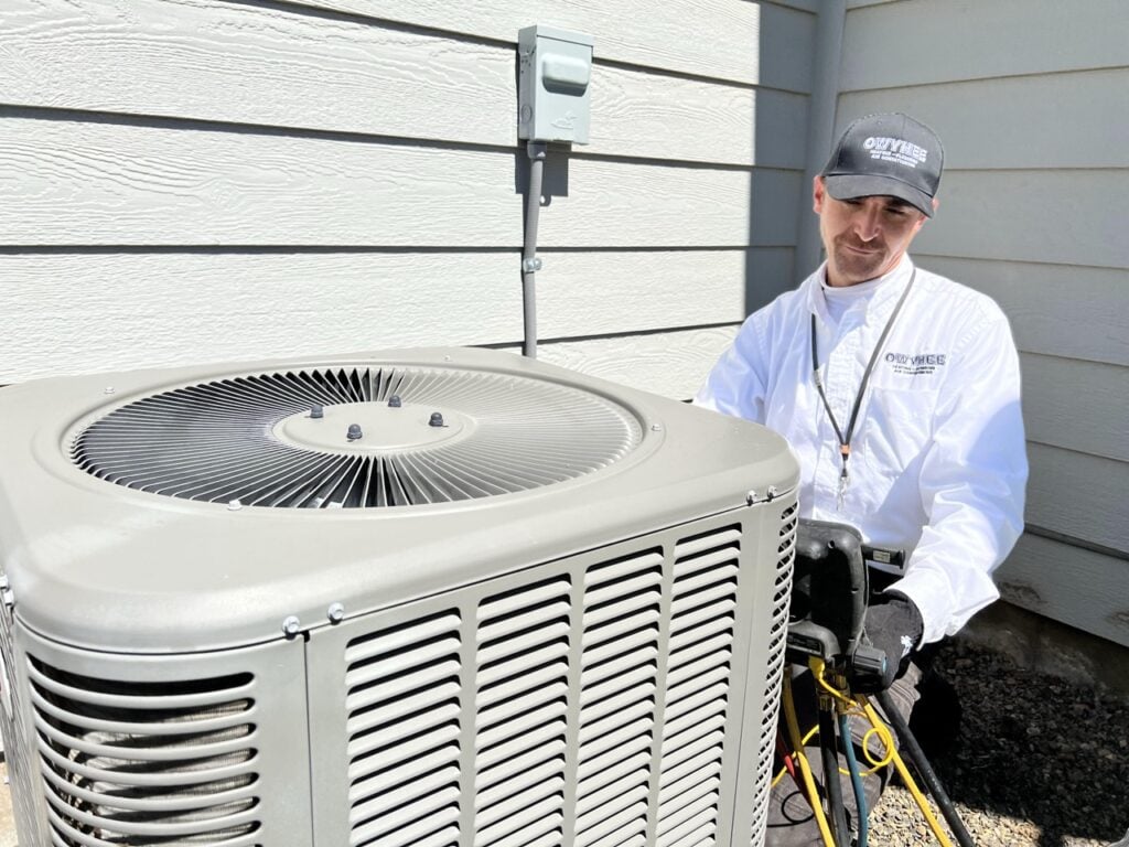 Professional technician in white shirt and cap standing beside a residential air conditioning condenser unit against a house with light-colored siding.