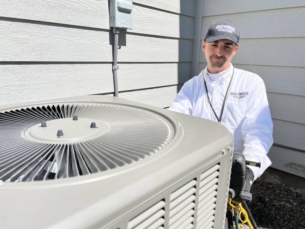 Professional technician in white shirt and cap standing beside a residential air conditioning unit outside a home.