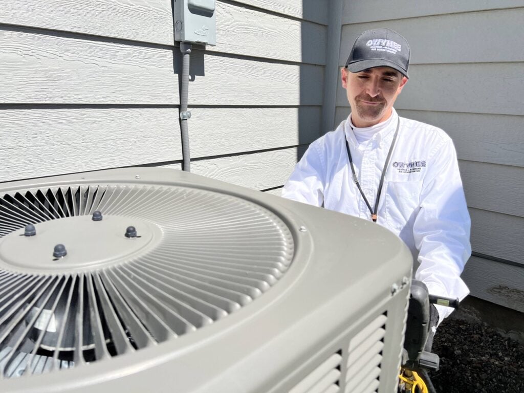 Professional HVAC technician in white shirt and cap stands beside a large air conditioning condenser unit outside a residential home.