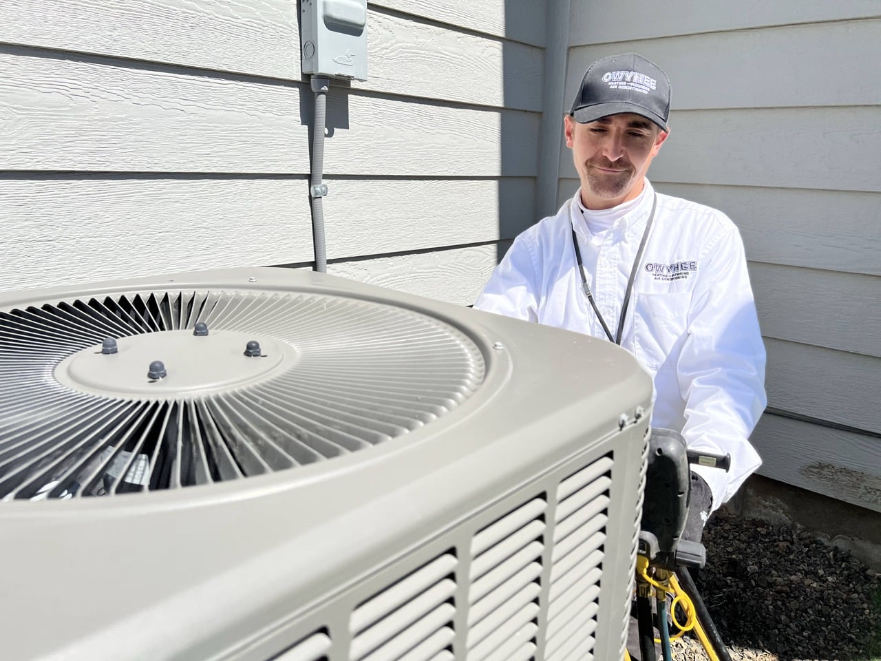 Professional HVAC technician in white shirt and cap posing next to a large outdoor air conditioning condenser unit against a house with fiber cement siding.