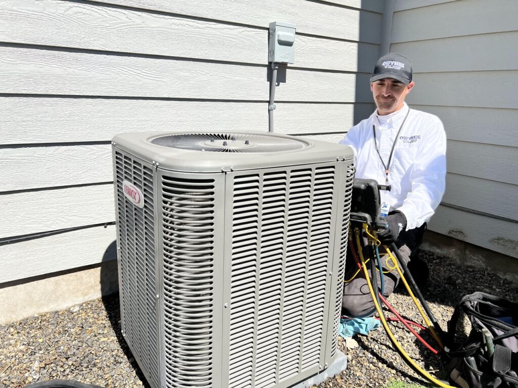 Professional HVAC technician in white shirt and cap standing beside a Lennox air conditioning condenser unit during installation at residential property.