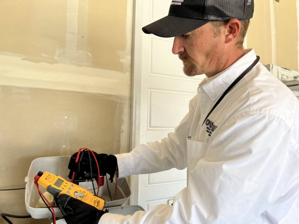 Electrician in white shirt and black cap uses a yellow multimeter to test an electrical outlet on a beige wall.