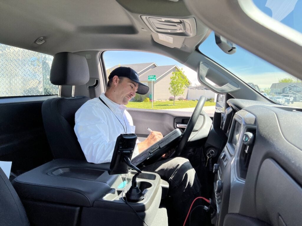 Man wearing light blue shirt and dark cap sits in driver's seat holding tablet, suburban neighborhood visible through windshield.