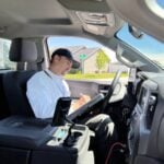 Man wearing light blue shirt and dark cap sits in driver's seat holding tablet, suburban neighborhood visible through windshield.