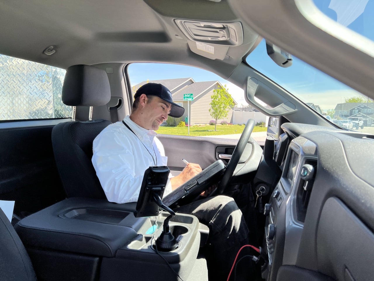Man wearing light blue shirt and dark cap sits in driver's seat holding tablet, suburban neighborhood visible through windshield.