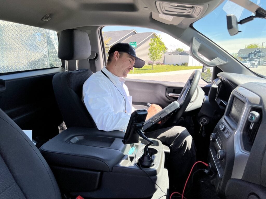 Man wearing light blue shirt and dark cap sits in driver's seat holding steering wheel, with residential homes and green lawn visible through windows.