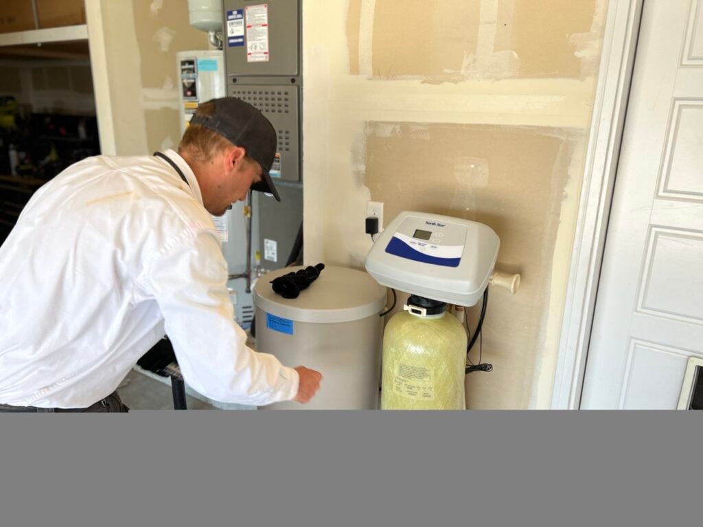 Technician in white jacket and black cap installing a North Star water softener system next to a residential water heater.