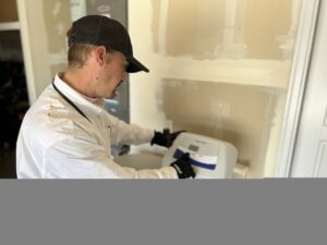 Technician in white jacket and black cap wearing gloves installs a digital thermostat on a beige wall.