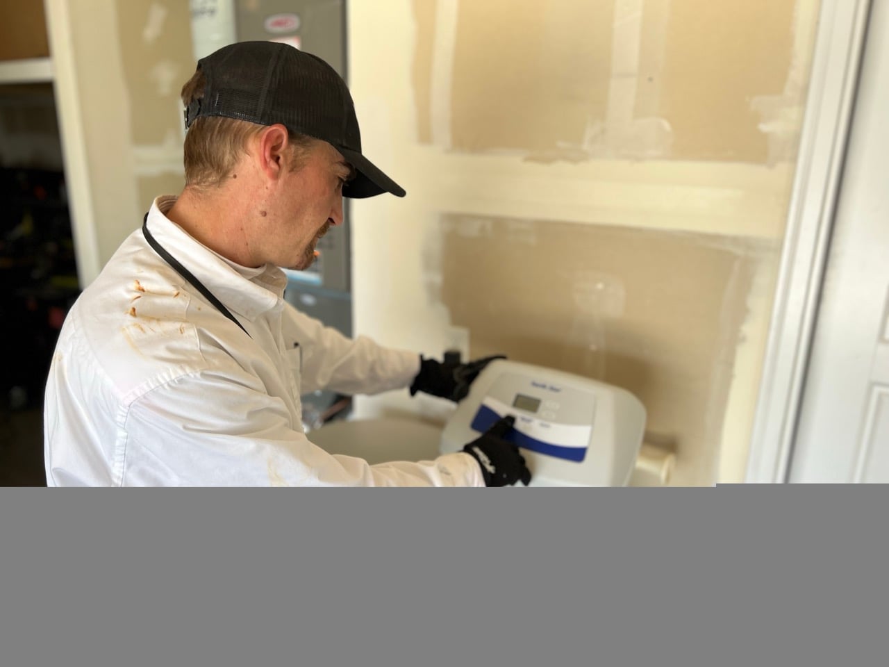 Technician in white jacket and black cap wearing gloves installs a digital thermostat on a beige wall.