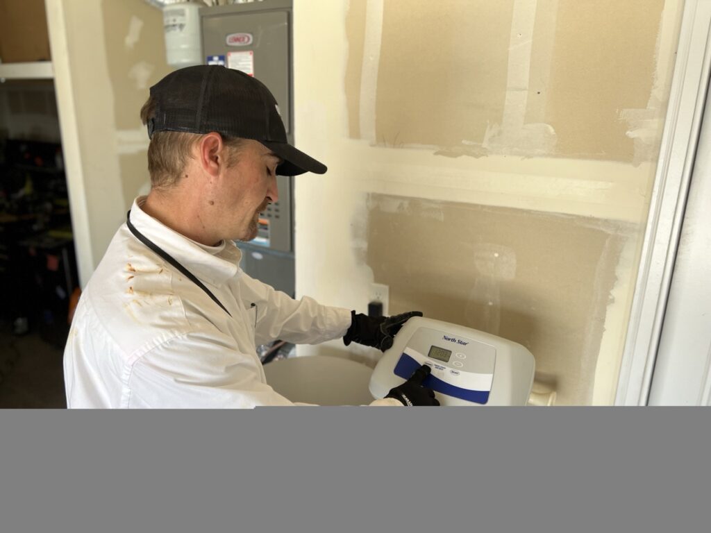 Technician in white shirt and black cap installs a North Star HVAC unit in drywall opening indoors.