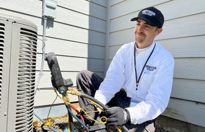 Professional HVAC technician in white shirt and cap uses diagnostic equipment on outdoor air conditioning condenser unit against white siding.